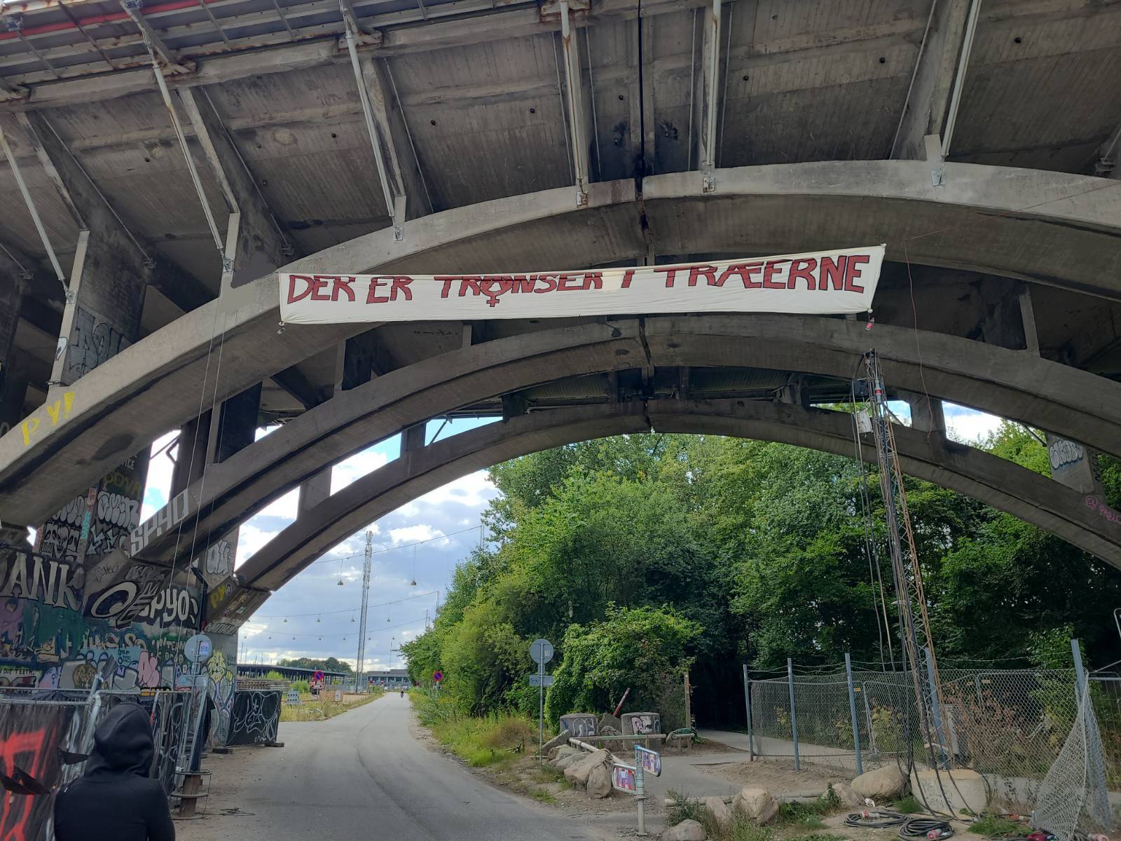A long banner on the side of a bridge with the text "Der er transer i træerne." or "There's trans folks in the trees"